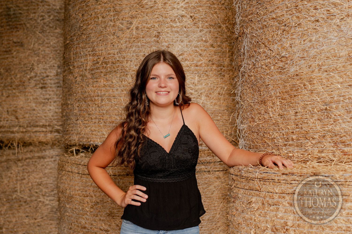 Portrait against round hay bales wearing black top and casual denim.