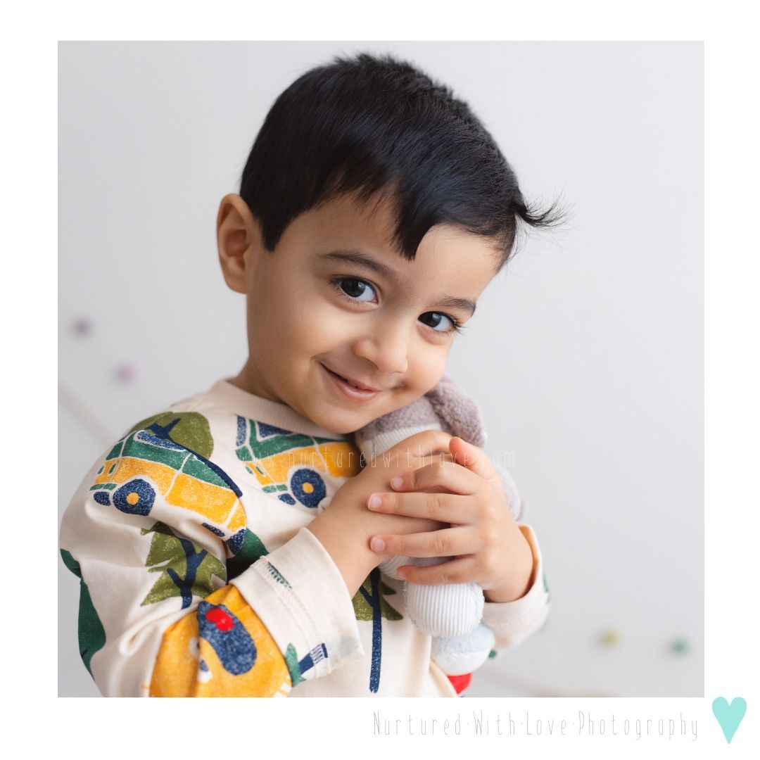 Child wearing a white sweatshirt with car and toucan print hugging a plush toy and smiling at the camera.
