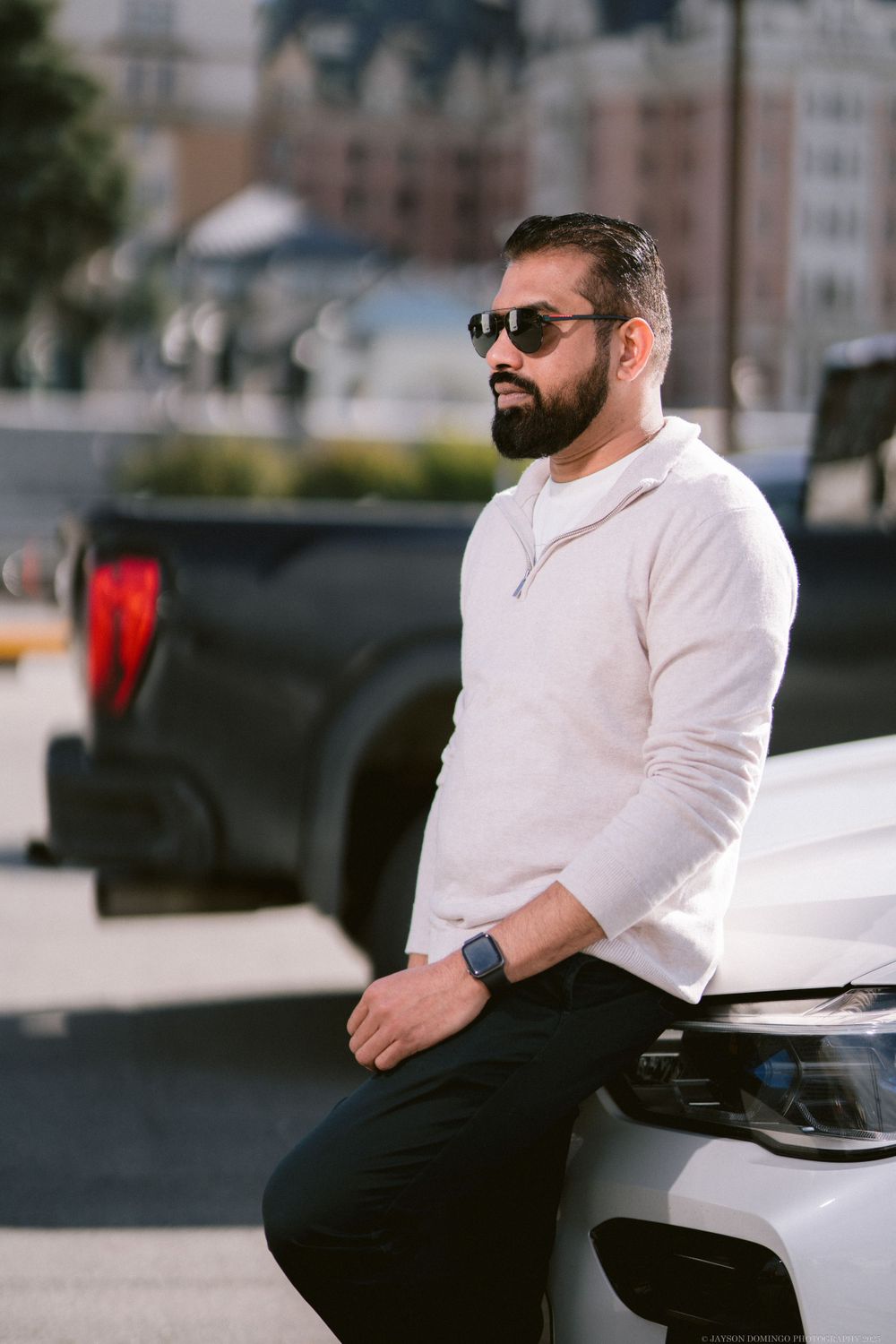 Casual portrait of someone in sunglasses and a white henley leaning against a white vehicle in an urban setting.