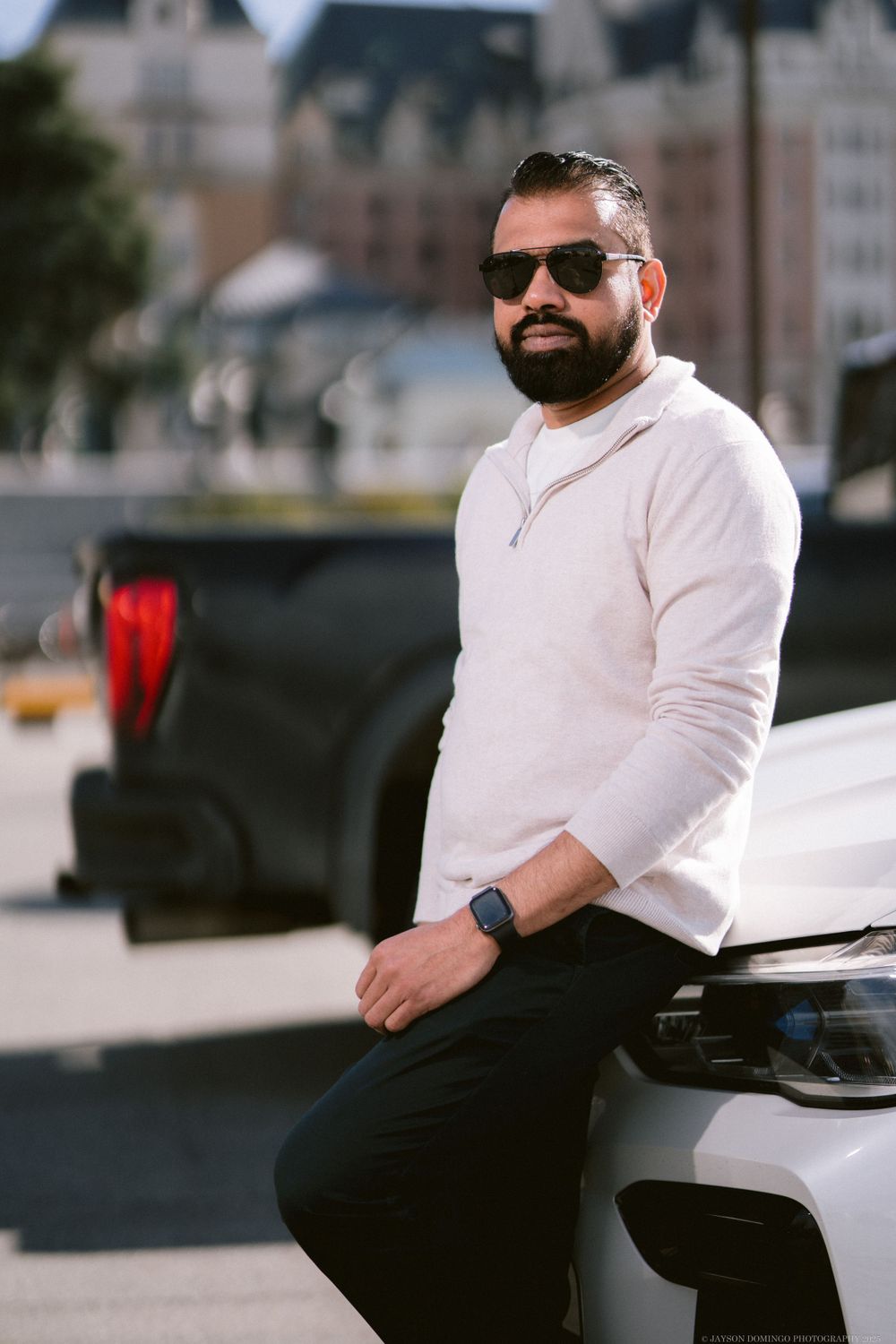A person in sunglasses and a cream sweater sits casually on a white car in an urban setting.