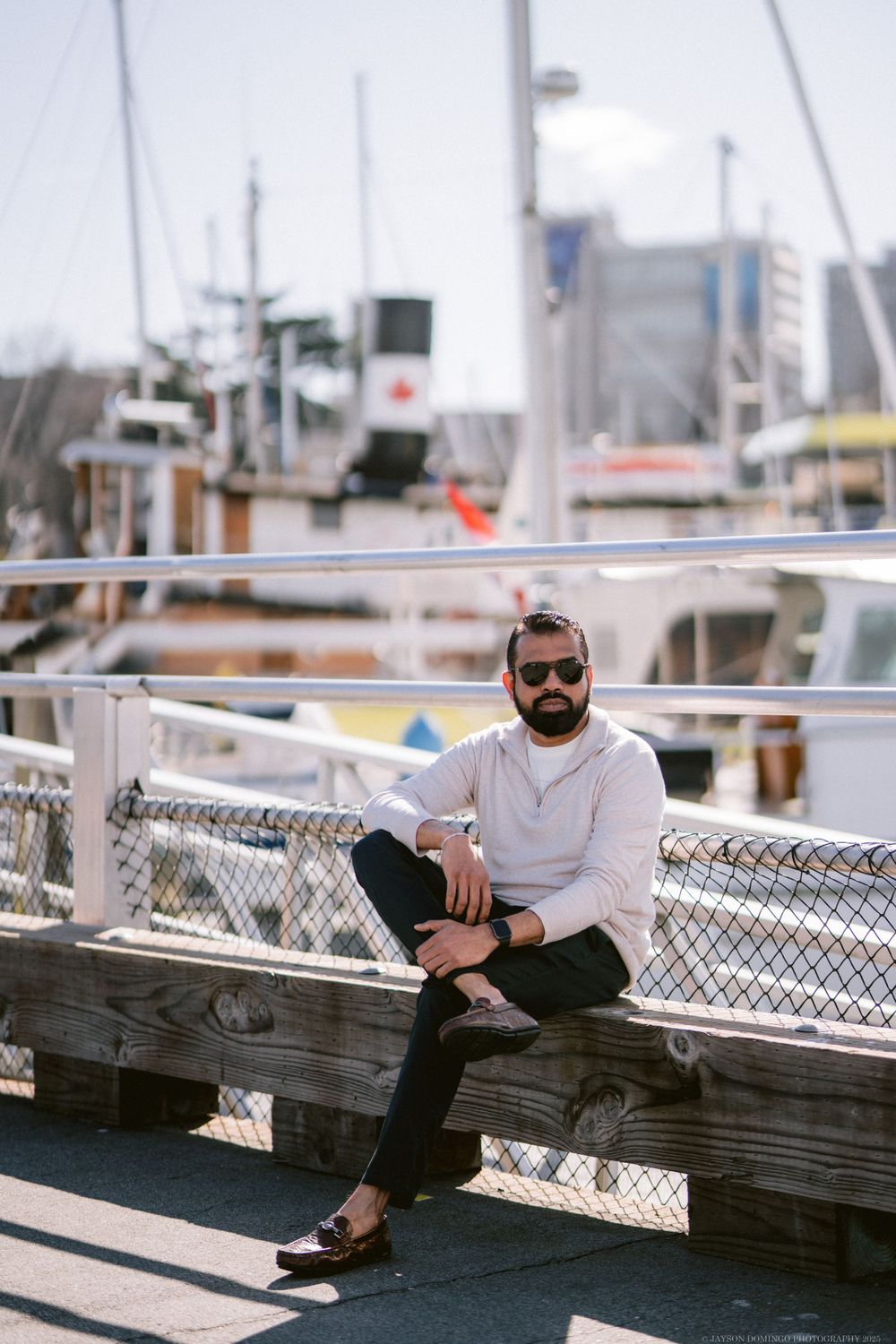 Casual figure wearing white hoodie and dark pants sits relaxed on wooden dock at marina with boats in background.