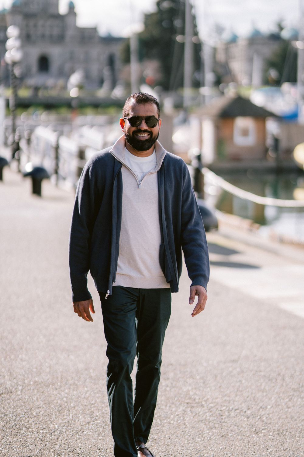 Casual pedestrian in sunglasses and navy jacket walking along waterfront path with castle-like buildings in background.