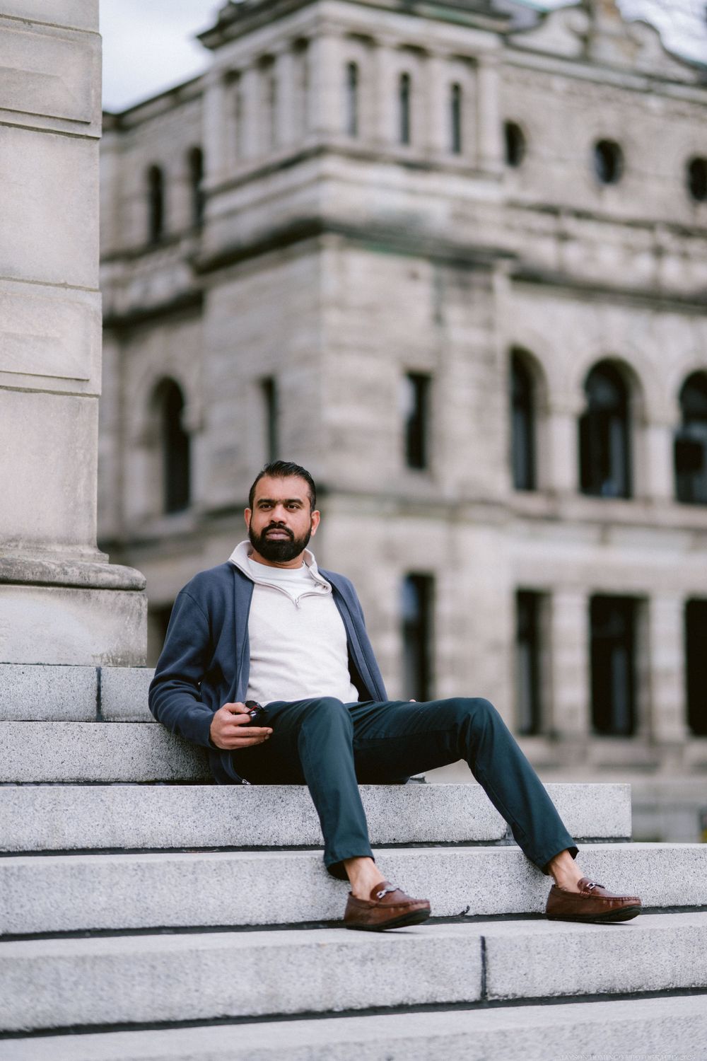 A person in casual attire sits on white marble steps of a classic architectural building while holding a phone.
