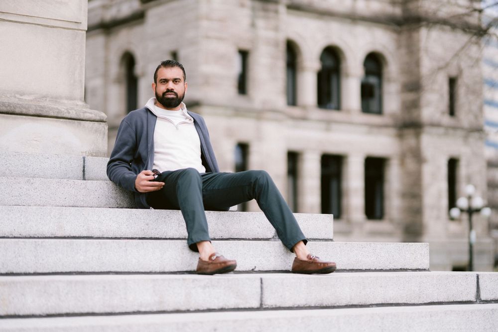 A casually dressed person sitting on stone steps holds a phone while wearing blue jeans and a gray blazer.