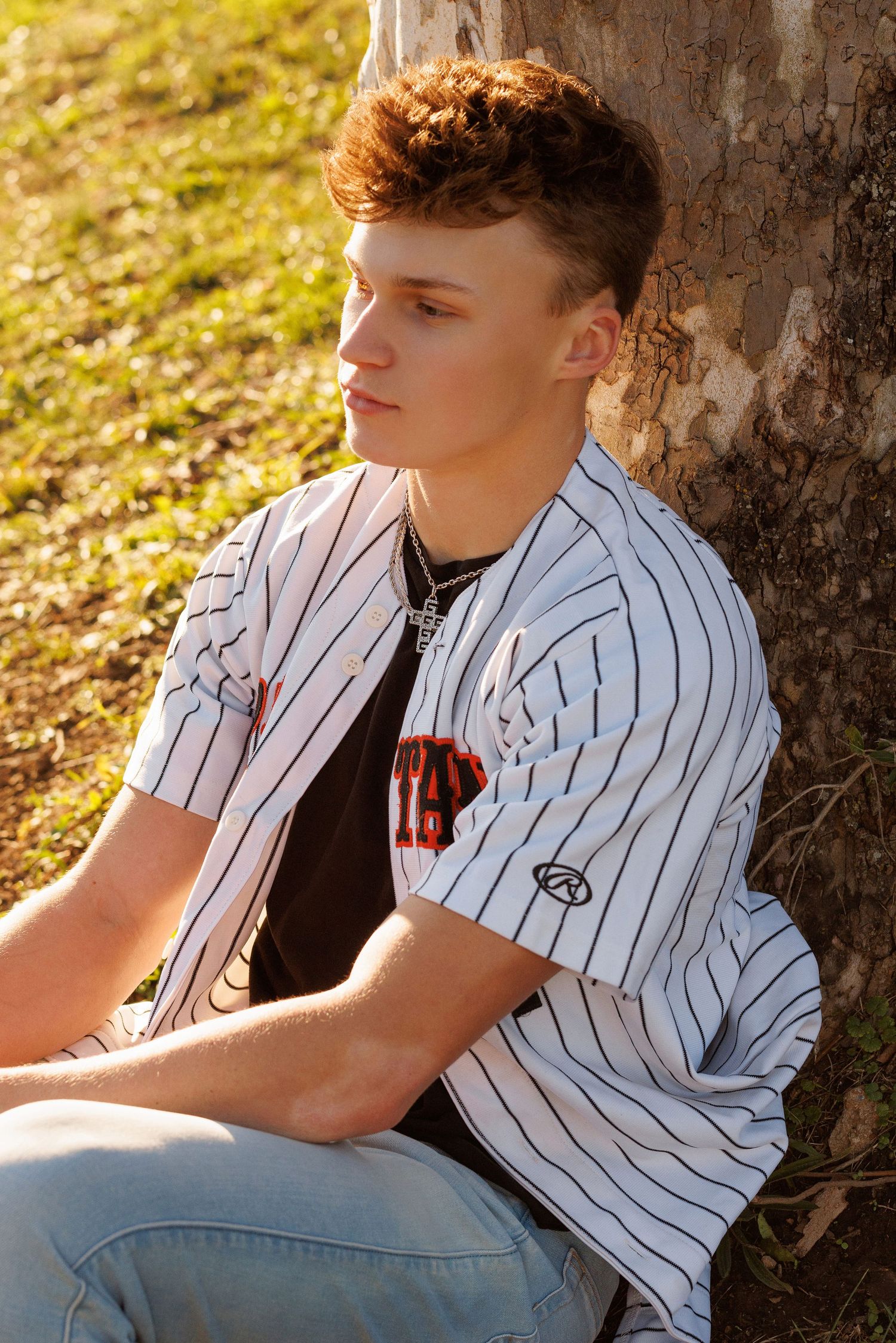 A person in a white pinstripe baseball jersey and jeans sits against a tree trunk in golden sunlight.