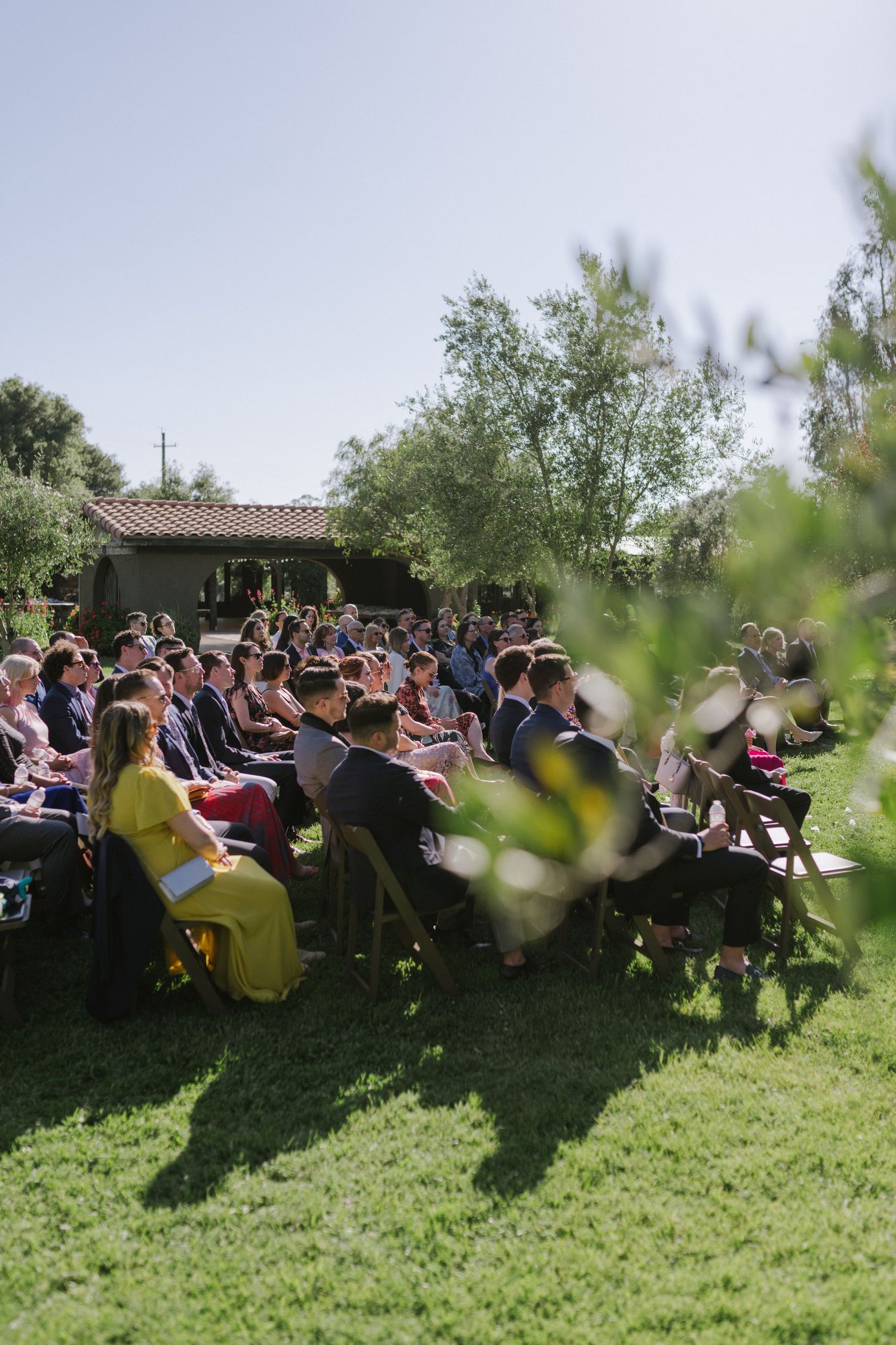 An outdoor wedding ceremony with guests seated on a sunny lawn near a Spanish-style building.