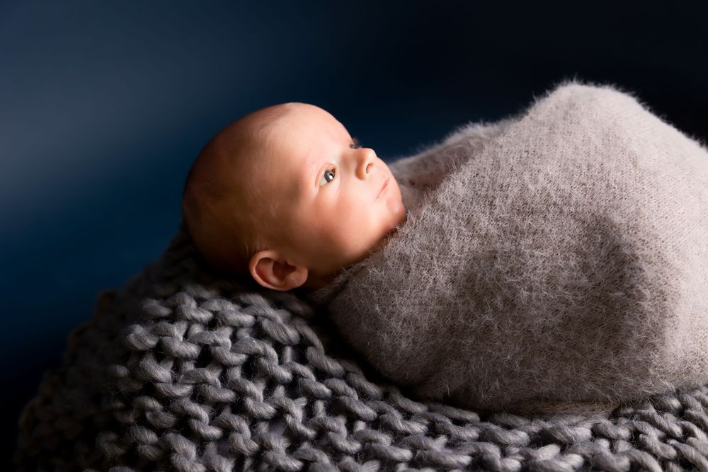 An infant nestled in a cozy gray knit blanket gazes upward against a dark navy background.