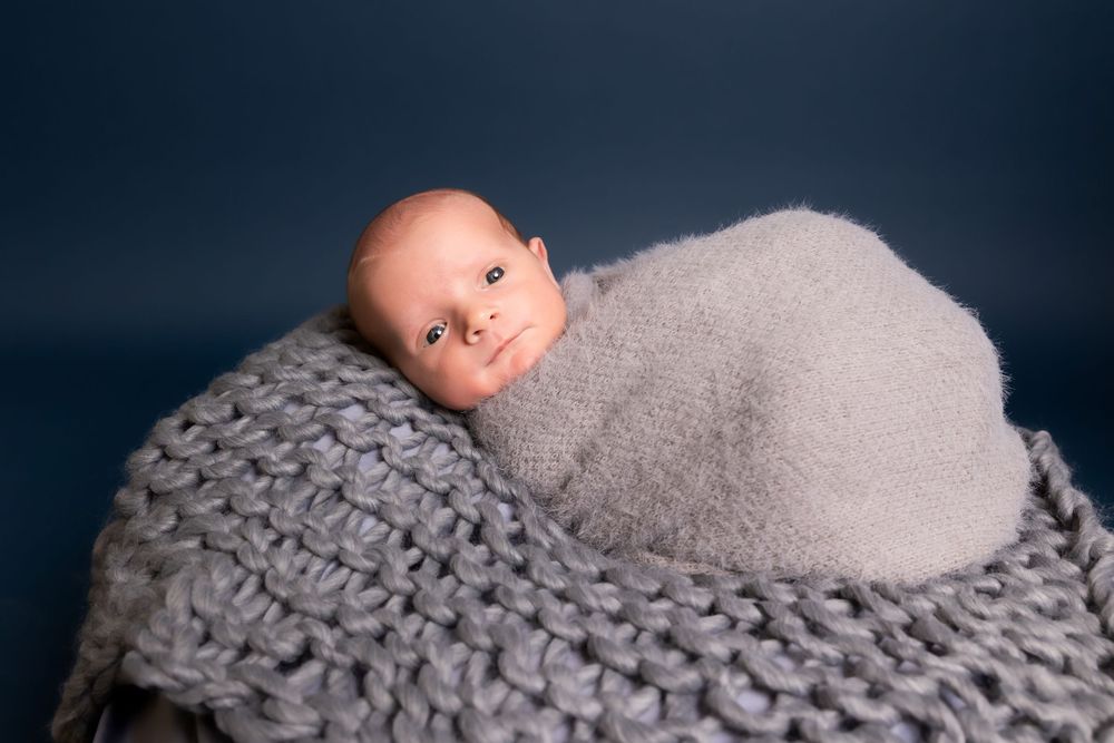 A newborn swaddled in soft gray fabric rests on a chunky knit blanket against a navy blue background.