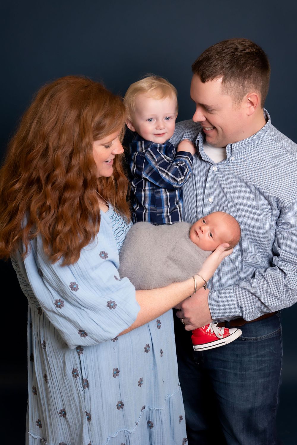 Family with young children poses in studio wearing coordinated blue and gray outfits against dark backdrop.