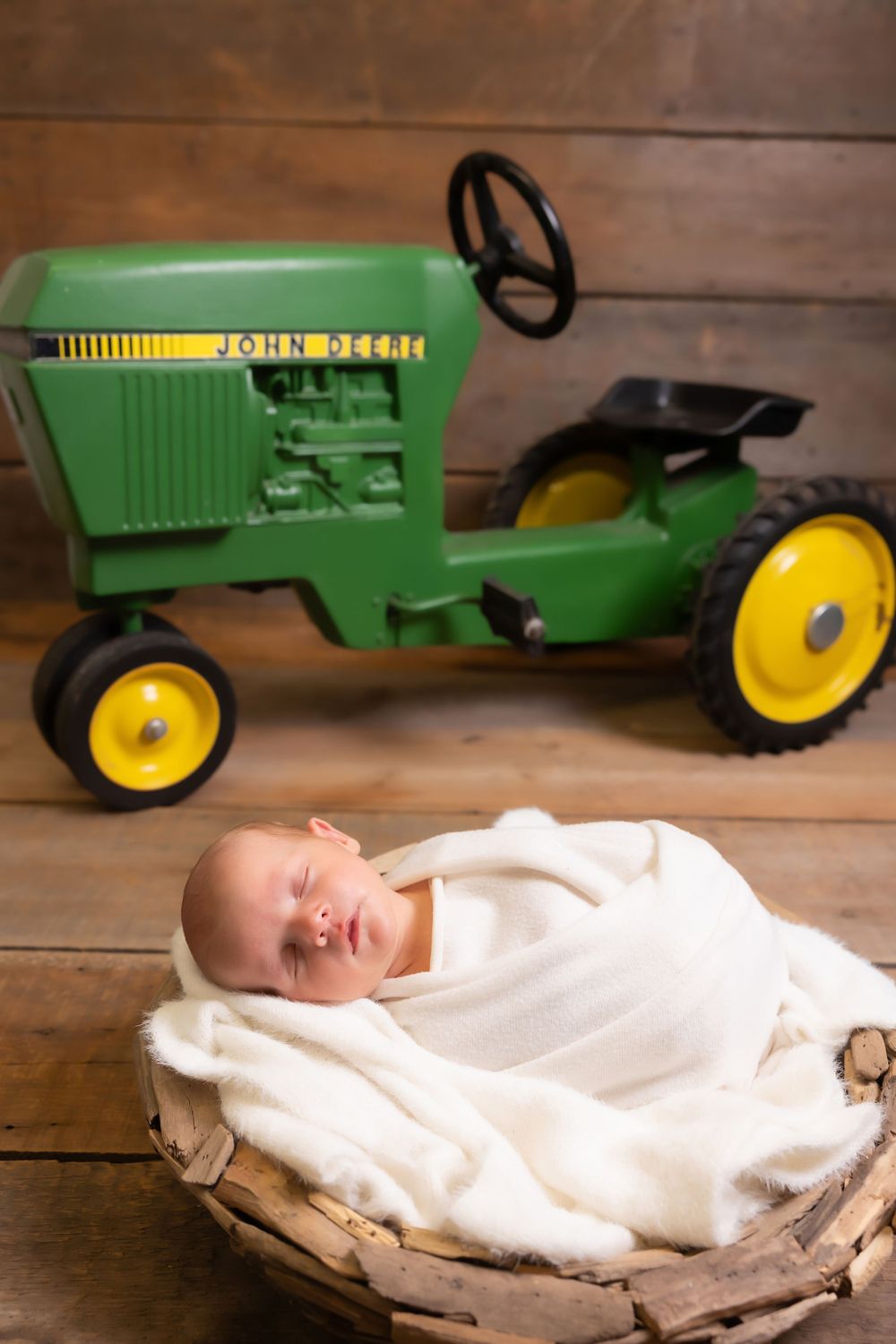 A sleeping newborn rests in a cozy white blanket basket next to a vintage green John Deere pedal tractor on wooden flooring.