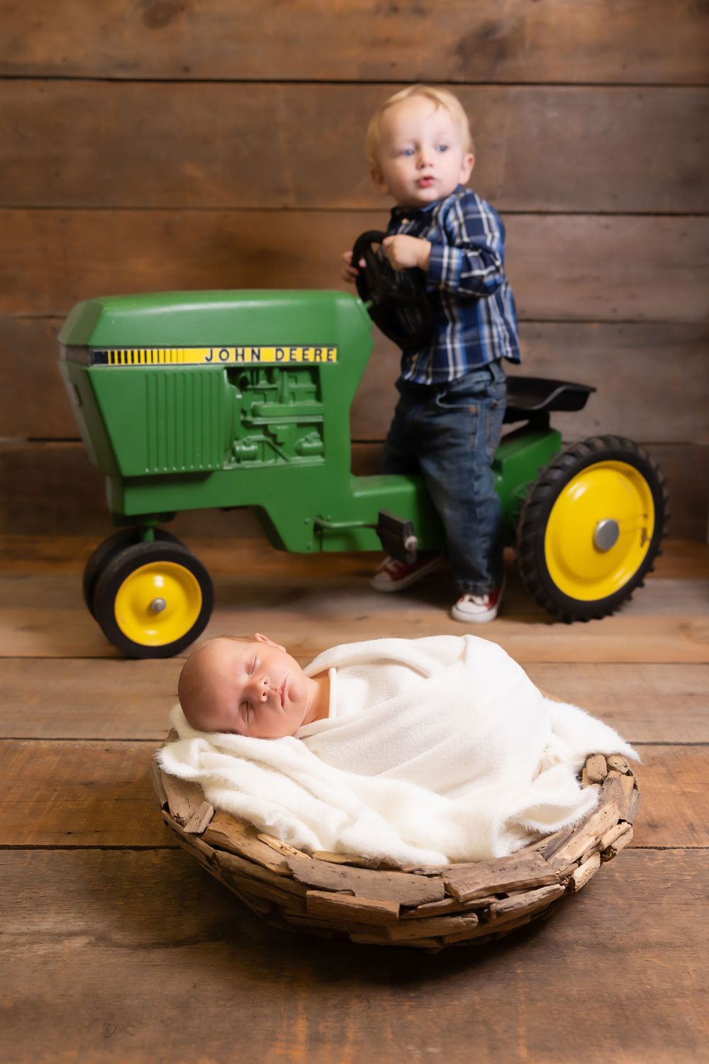 Two children pose beside a green John Deere toy pedal tractor on wooden flooring with one in a cozy basket.