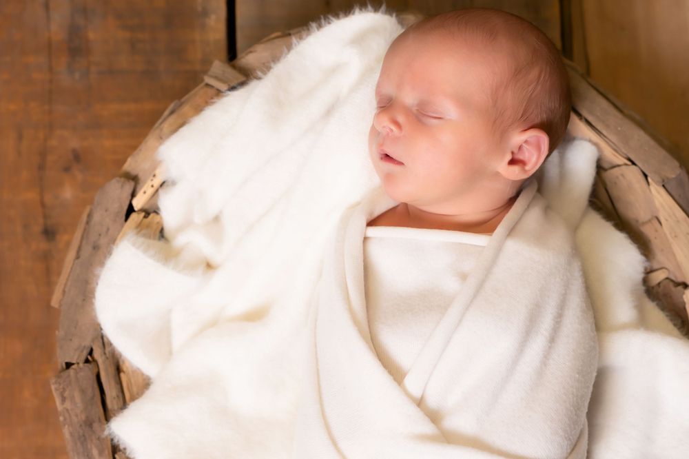 A sleeping newborn baby swaddled in white cloth lays peacefully in a wood basket.