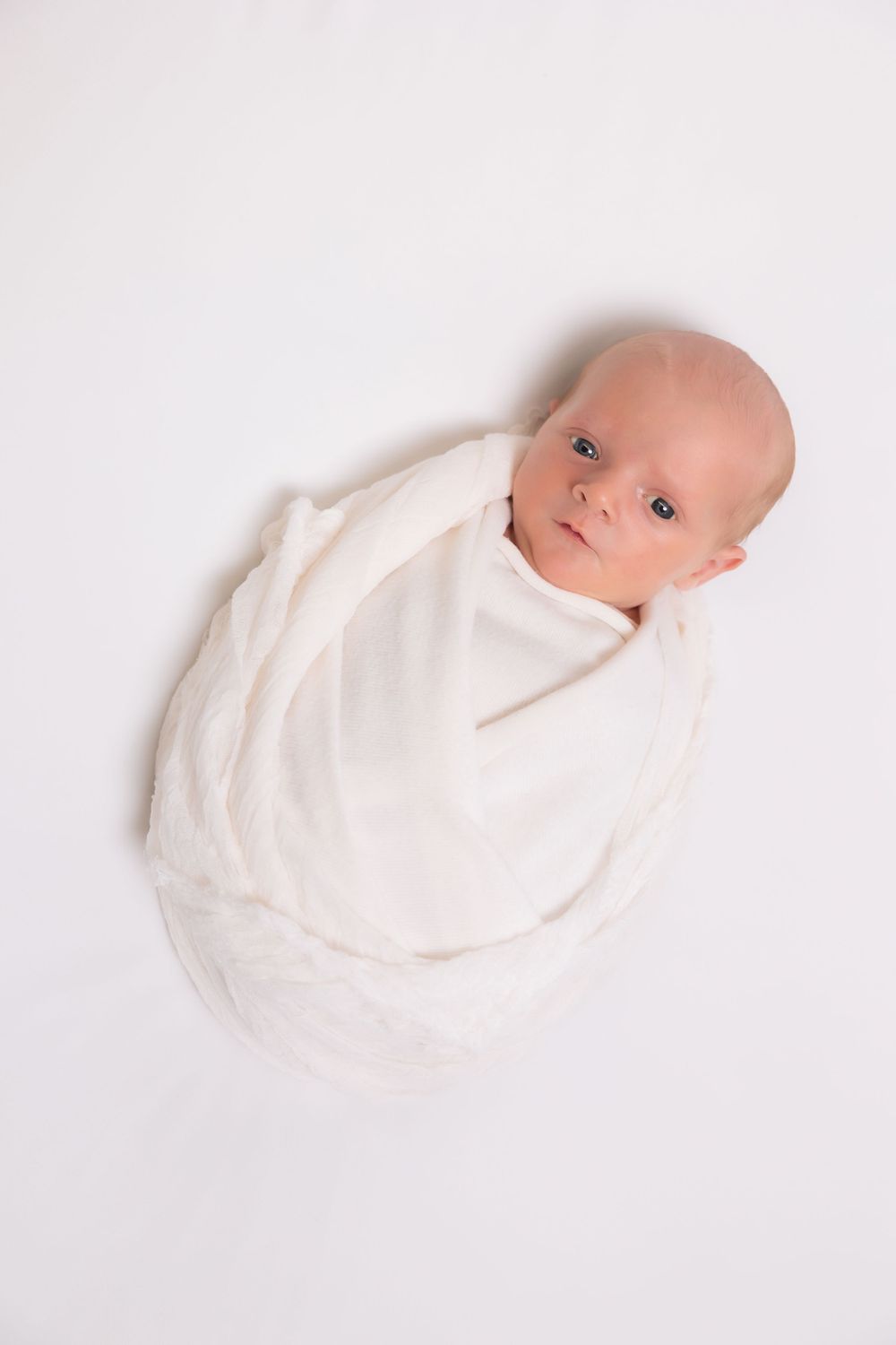 A baby swaddled in white fabric lies on a plain white background.