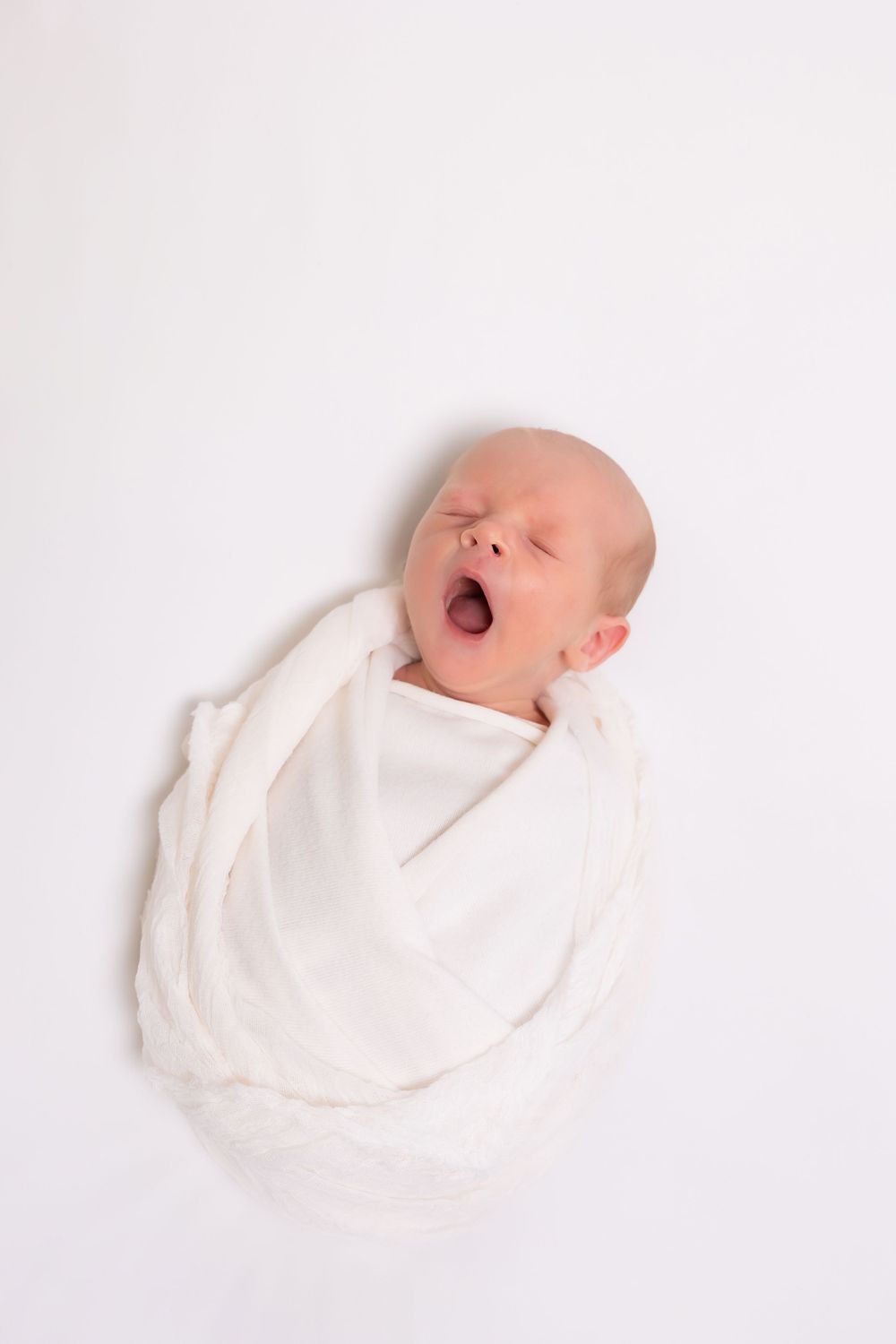 A newborn baby yawns while swaddled in a white blanket against a plain white background.
