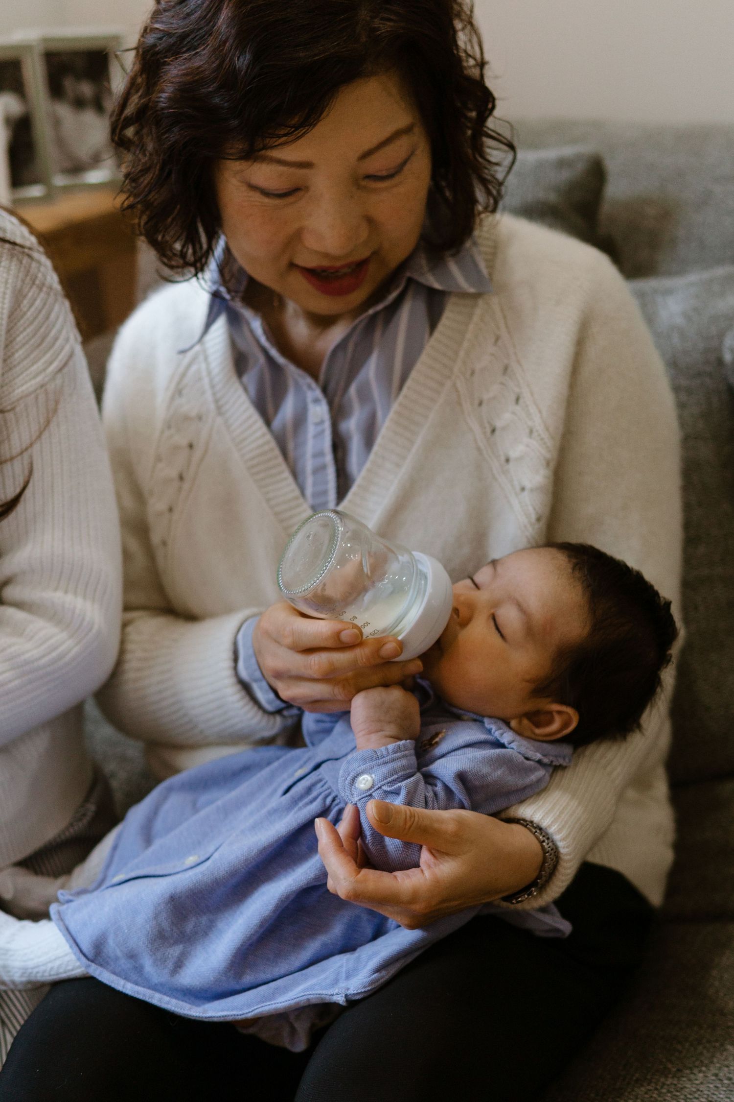 Baby in a blue outfit being bottle fed while resting on grandmother's lap on a couch.
