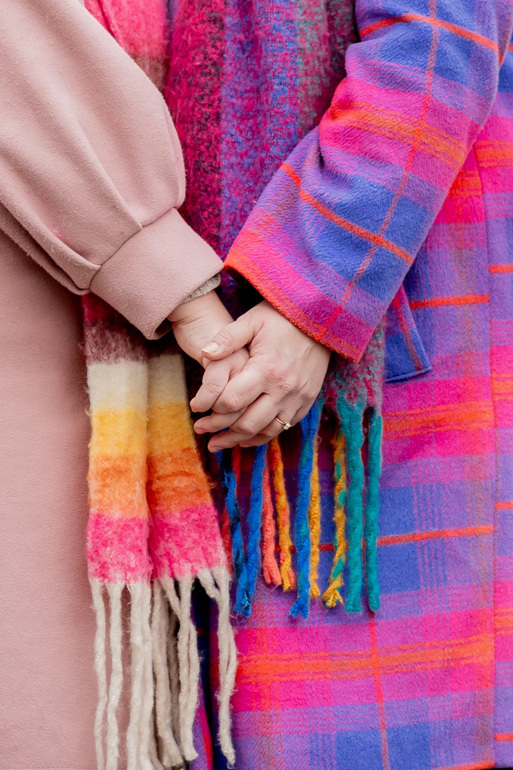 Close-up of hands holding while wearing vibrant rainbow scarf and pink coat against colorful textile background.