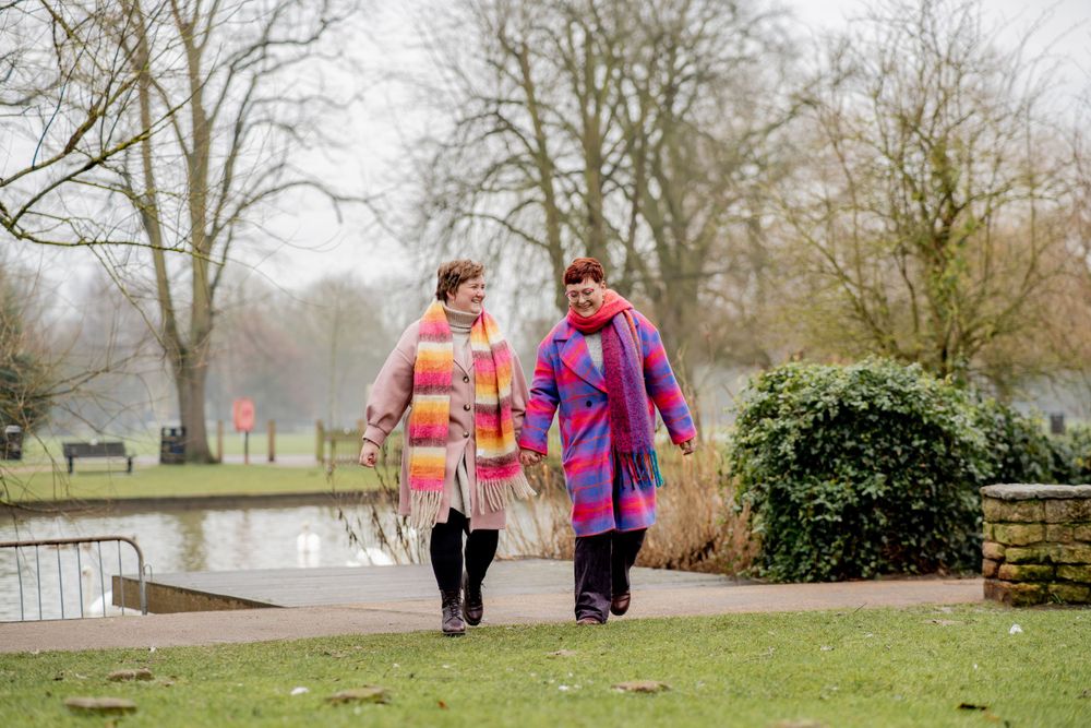 Two people in vibrant patterned coats walk together along a peaceful park path near a pond on a misty day.