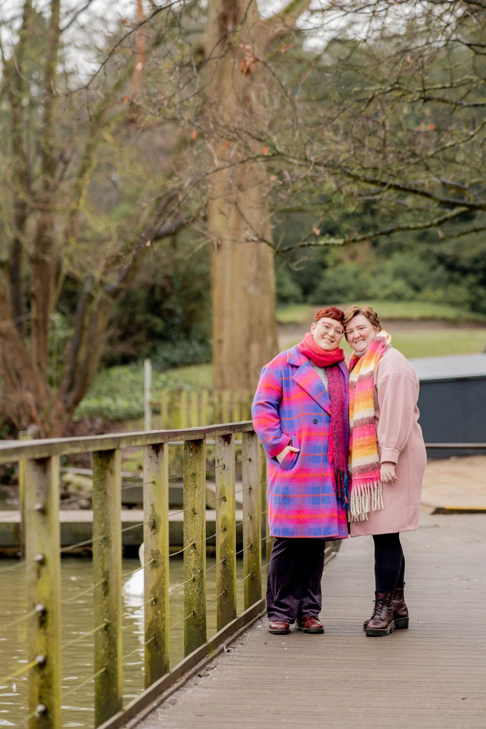 Two people in colorful winter coats standing together on a wooden boardwalk near a lake on a cloudy day.