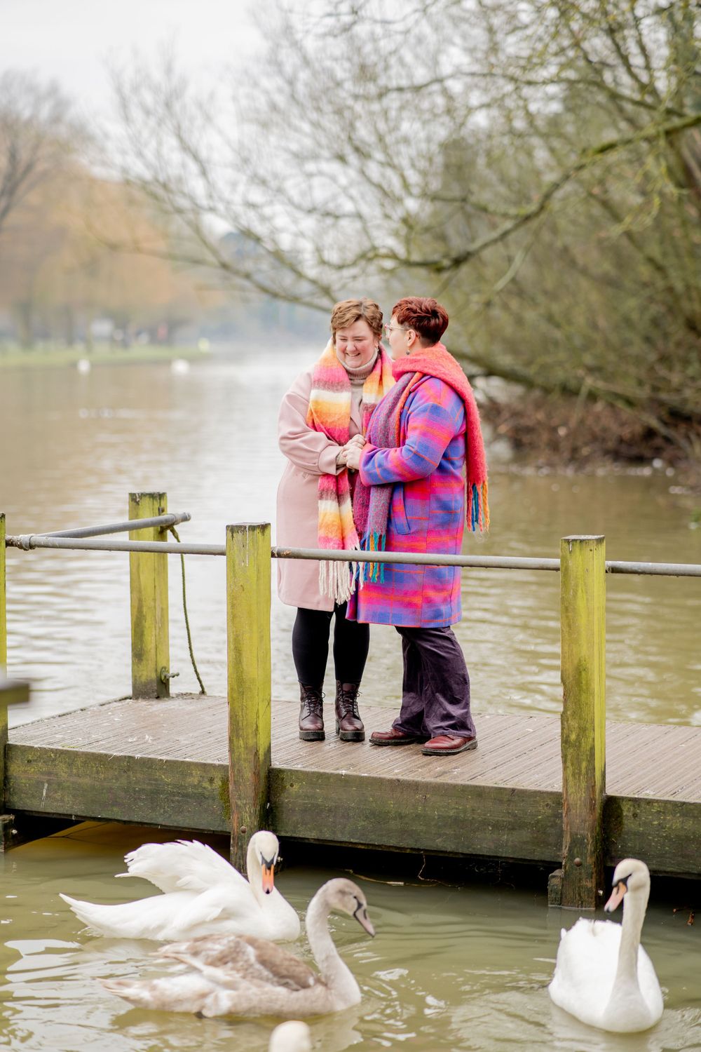 Two people in colorful winter coats stand on a wooden dock feeding swans at a lake on a misty day.