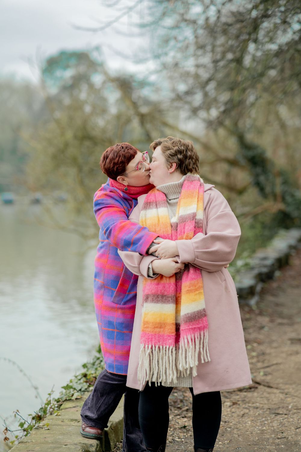Two people in winter coats share a tender moment by a lake on a misty day wearing colorful scarves.