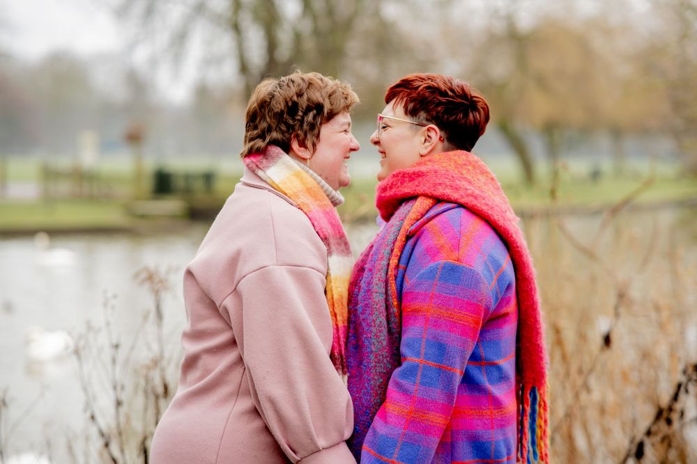 Two people in warm winter coats and scarves share a tender moment by a lake on an autumn day.