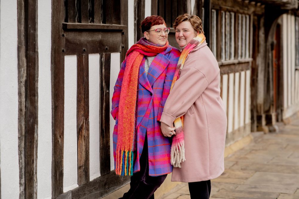 A couple in winter coats sharing an intimate moment against a historic Tudor-style building backdrop.
