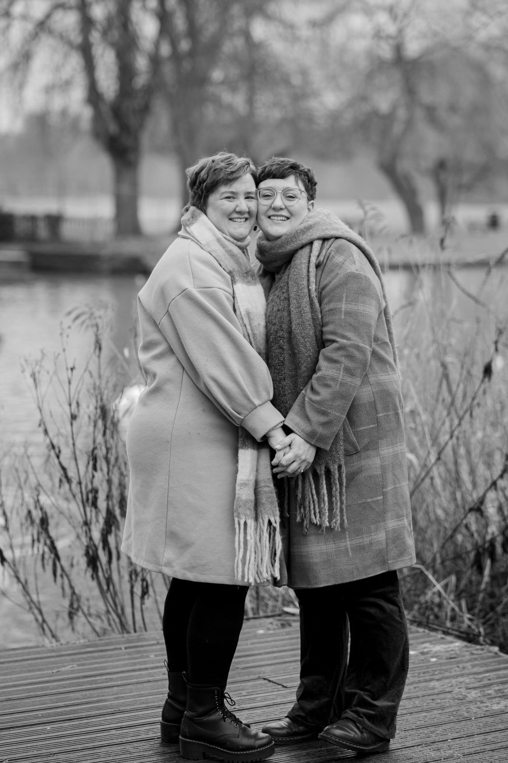 A romantic black and white portrait of a couple in winter coats embracing on a wooden dock.