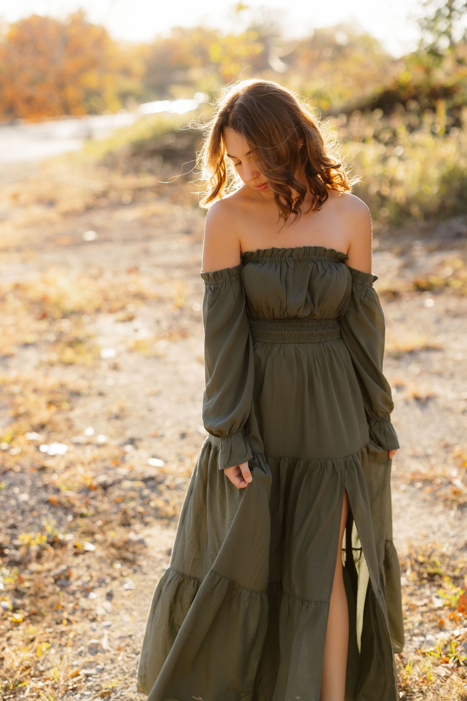 Woman in olive green off-shoulder maxi dress poses outdoors during golden hour sunset in autumn setting.