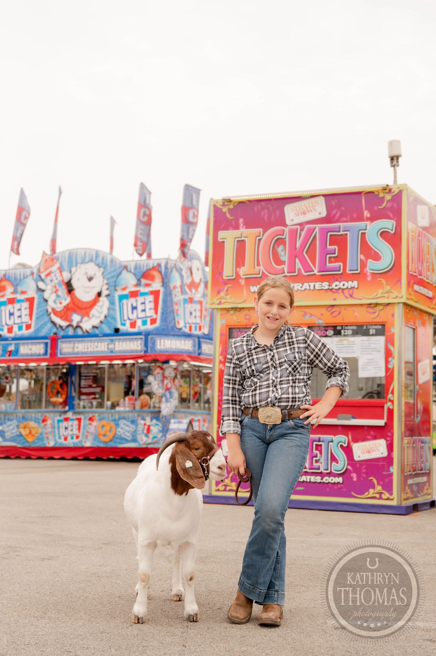 Goat and handler pose together by ticket booth at carnival with colorful game stalls in background.