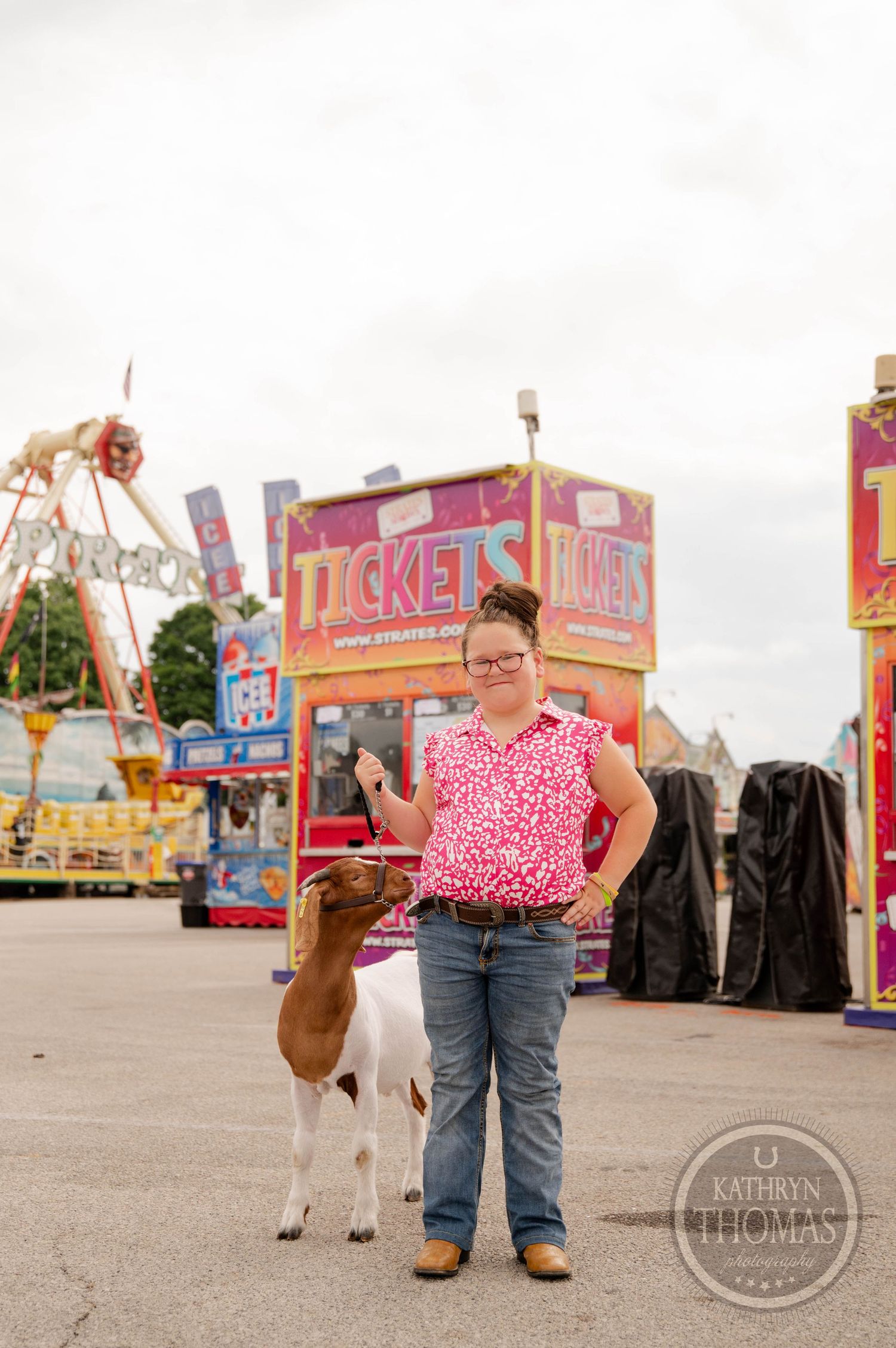 A brown and white goat stands near carnival rides and ticket booths at a fairground.