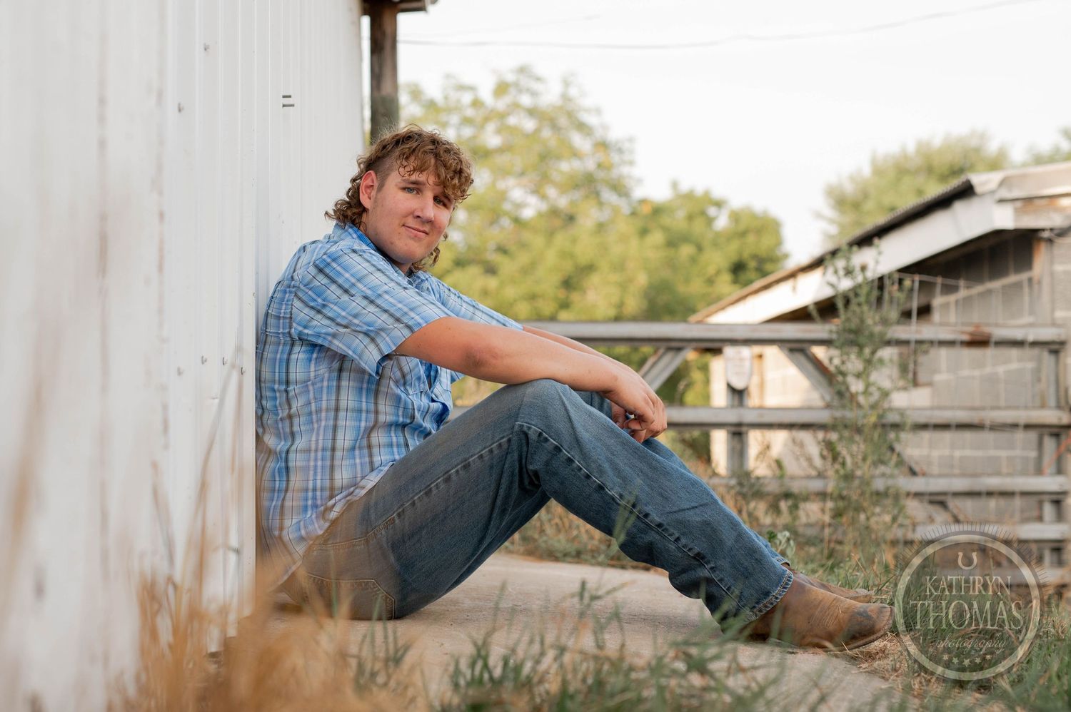 A relaxed pose against a barn wall wearing a blue plaid shirt and jeans.