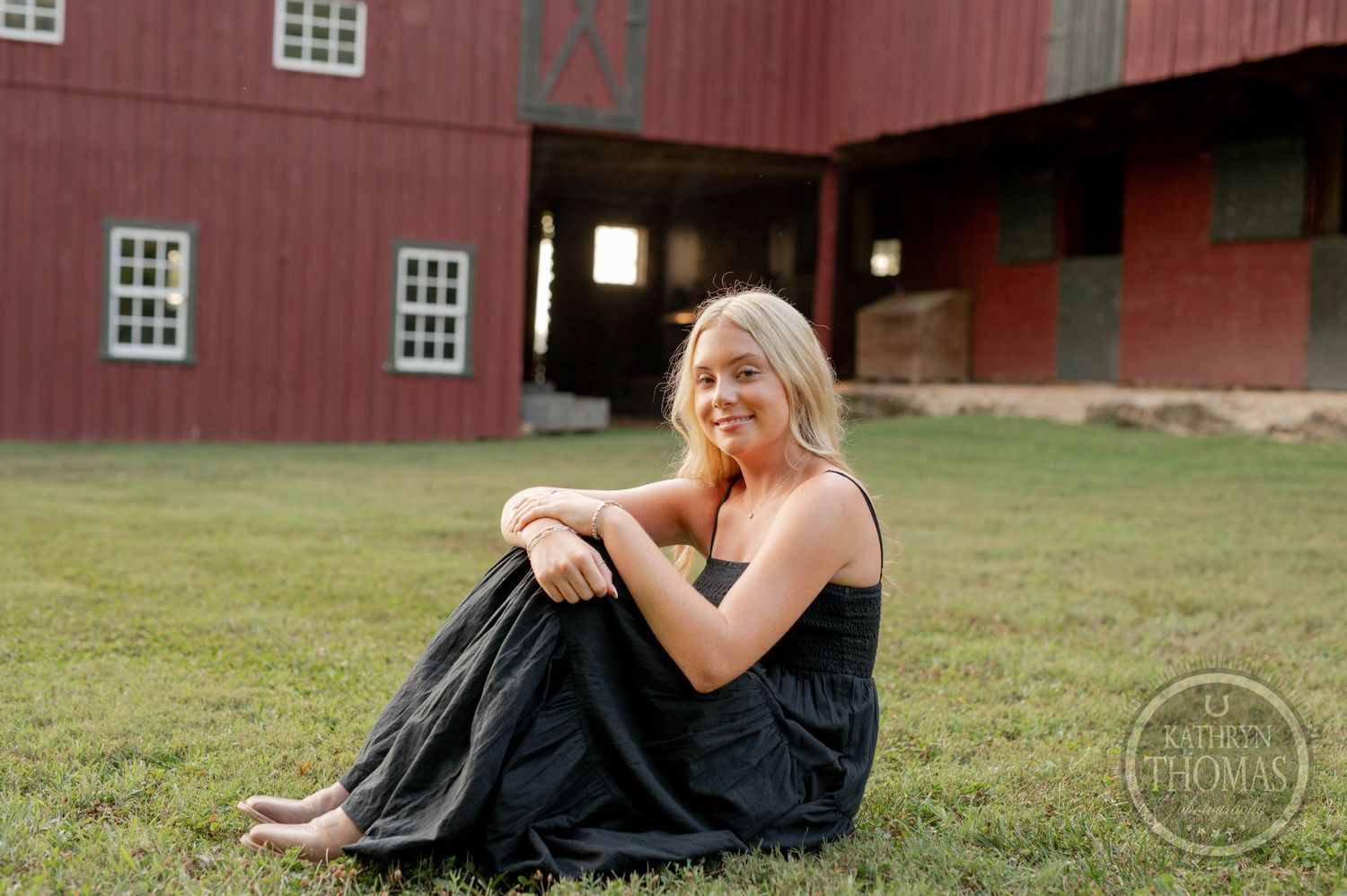 A woman sits on grass wearing a black dress with a historic red barn in the background.
