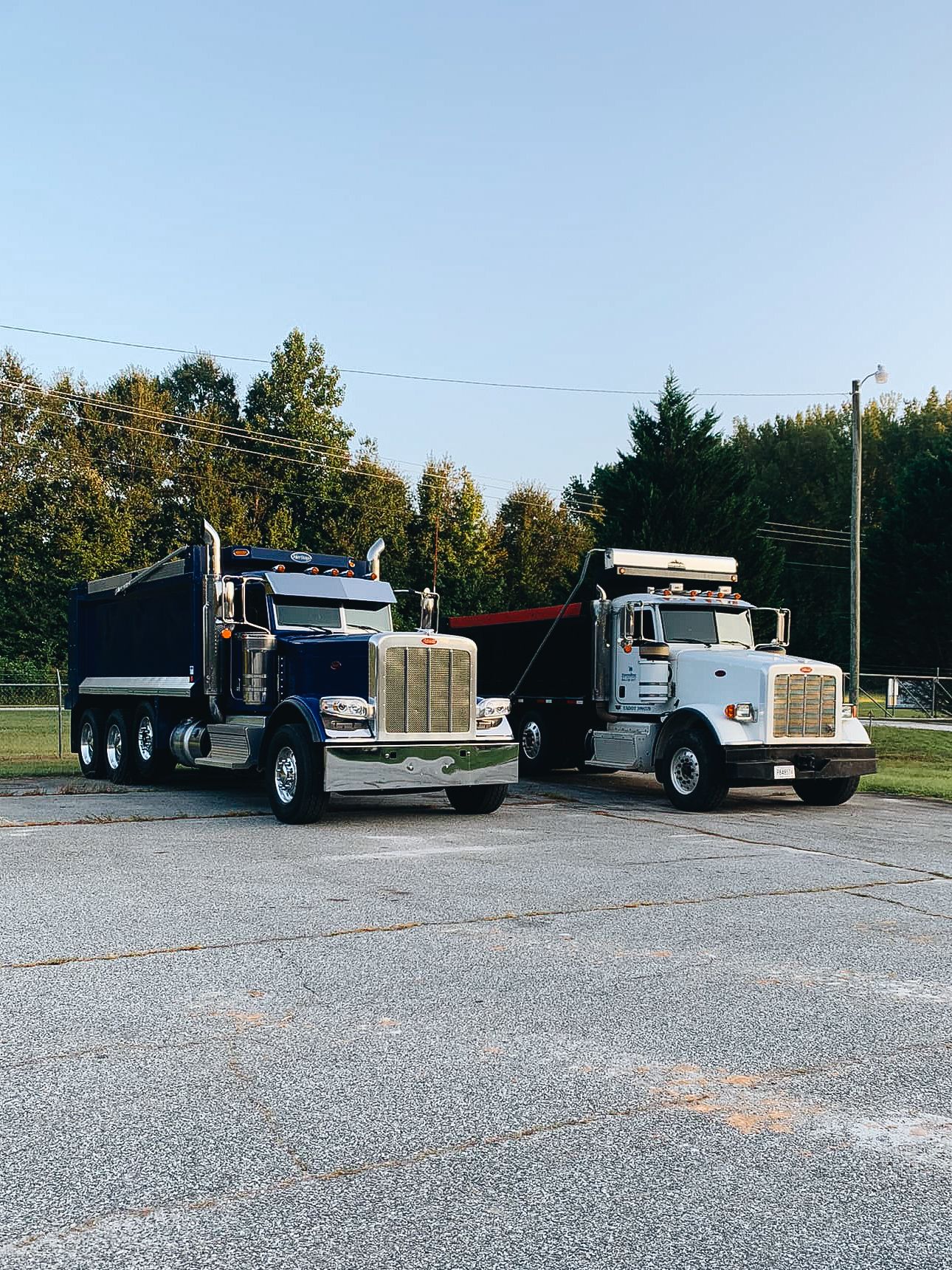 Two large dump trucks parked side by side in a lot with blue sky overhead.