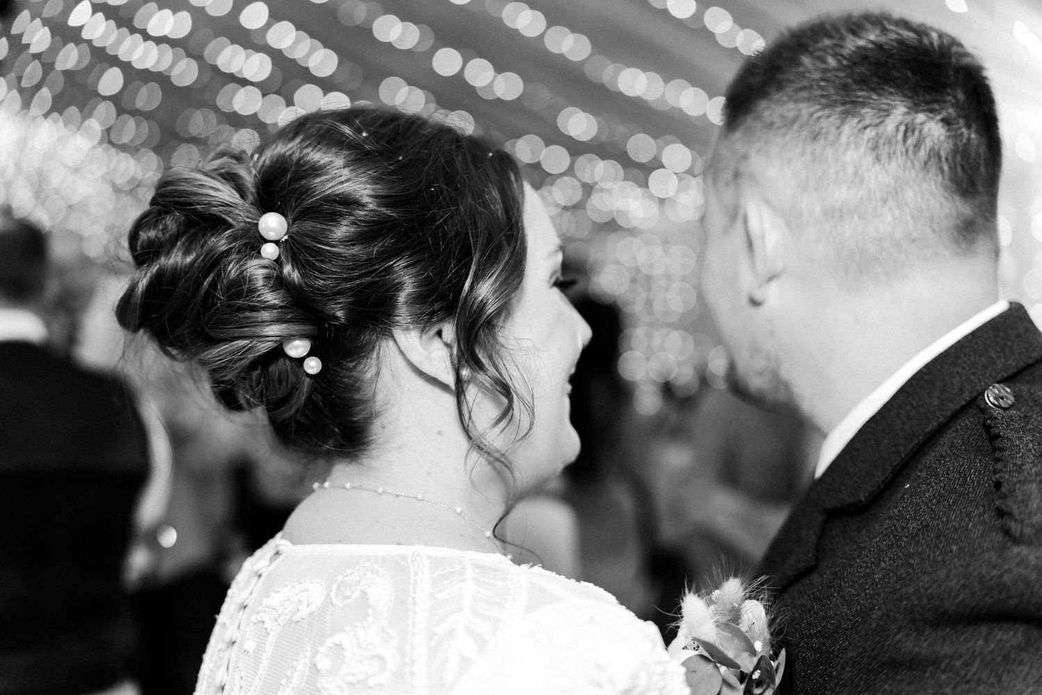 Black and white photo showing an elegant updo hairstyle with delicate pearls at a wedding reception with bokeh lights.