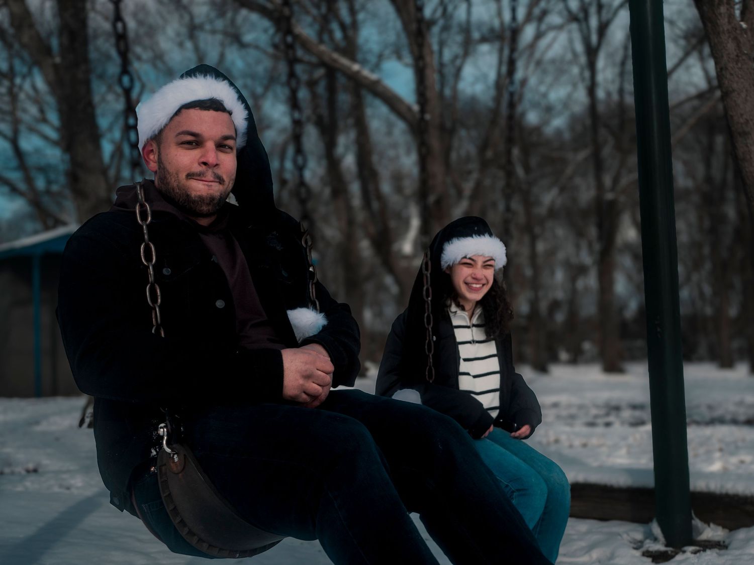 Two people wearing winter hats sit together on a snowy swing in a wooded setting at dusk.