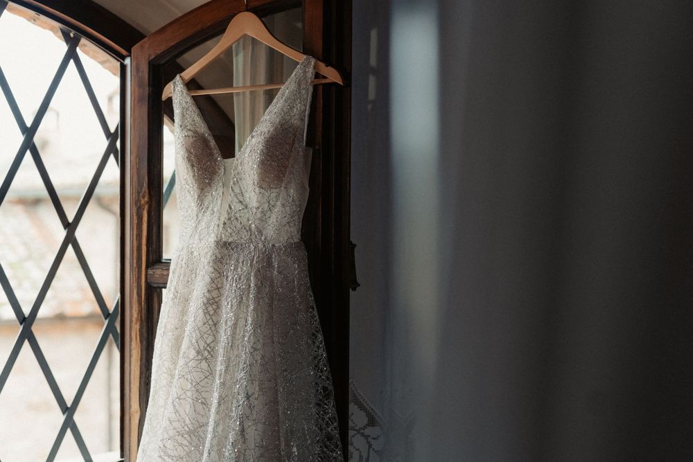 A delicate lace wedding dress hangs elegantly by a window with diamond-patterned glass panes.