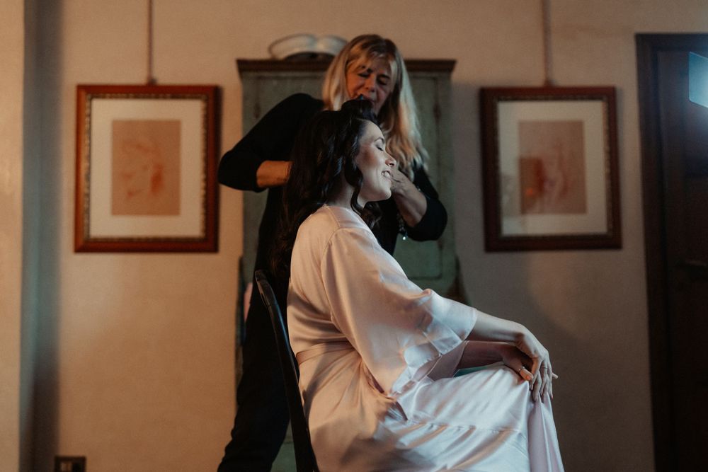 Stylist in black attends to someone wearing white during wedding preparations in a vintage-styled room.