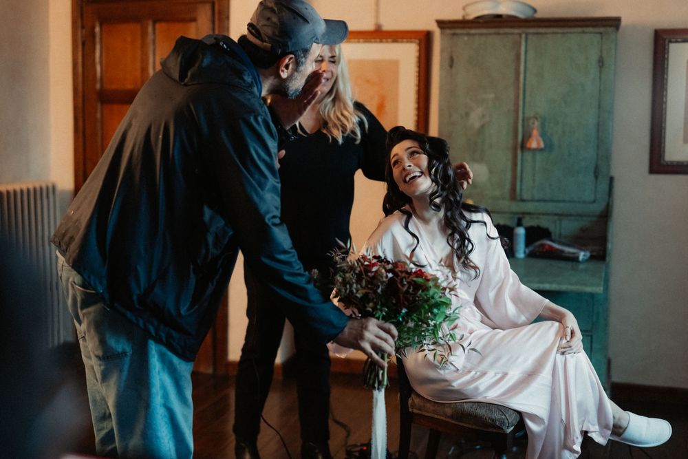 Two people helping with wedding preparations as someone in white holds a bouquet of flowers.