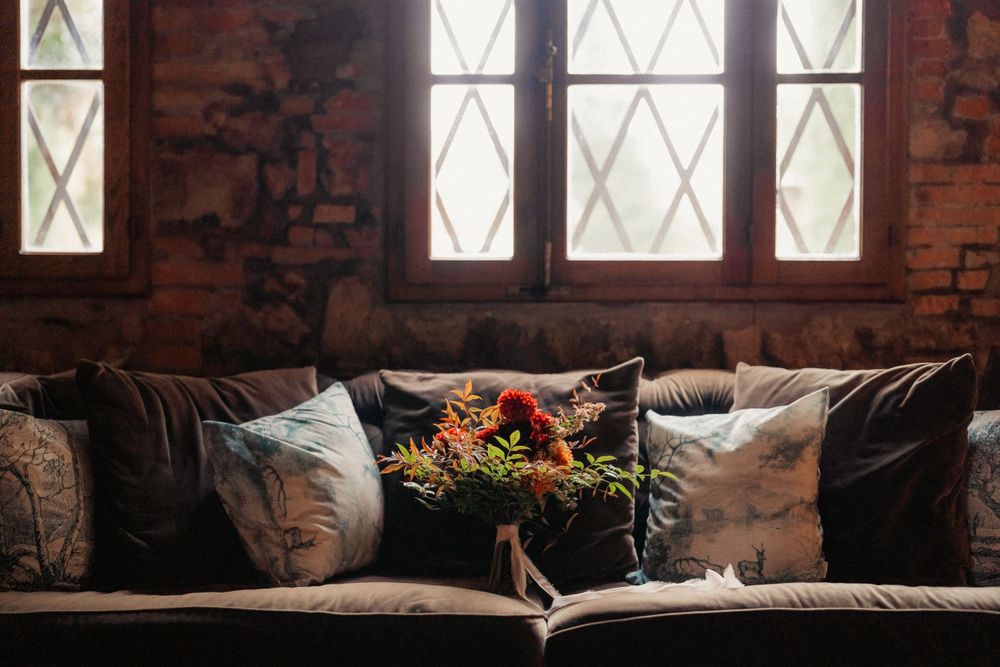 A cozy leather couch with throw pillows and floral centerpiece near vintage windows in a rustic brick interior.