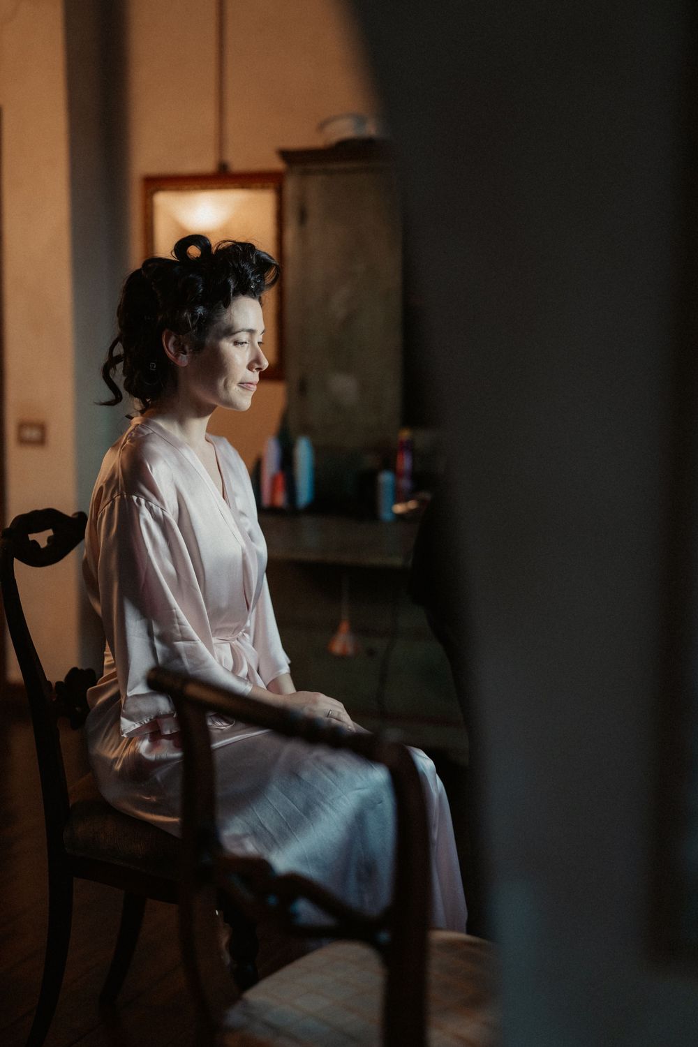 A person in white satin robe sits contemplatively in a chair with moody side lighting through a window.