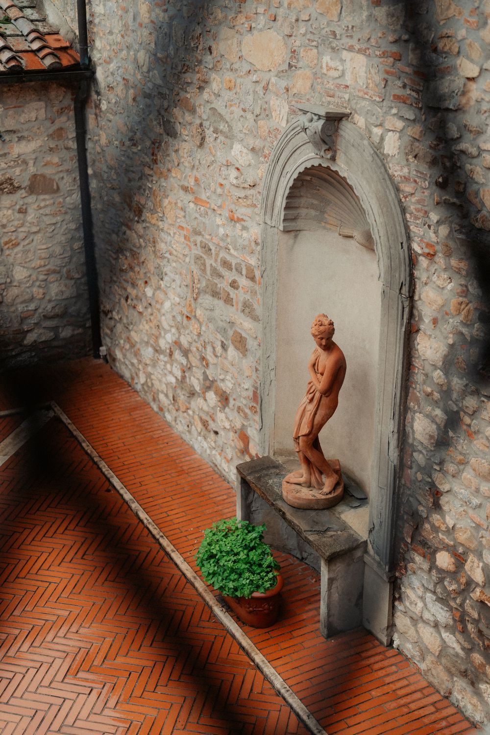 A rustic stone archway with a terracotta brick walkway featuring a classical nude statue and small potted plant.