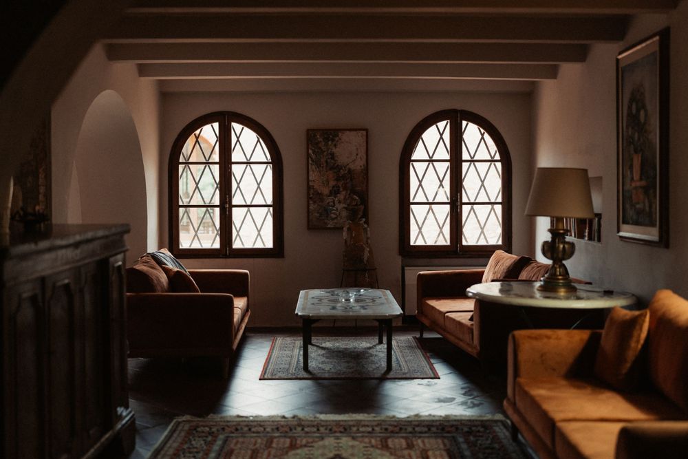 A cozy vintage living room features gothic arched windows, brown leather couches, and dark wood paneling in warm light.