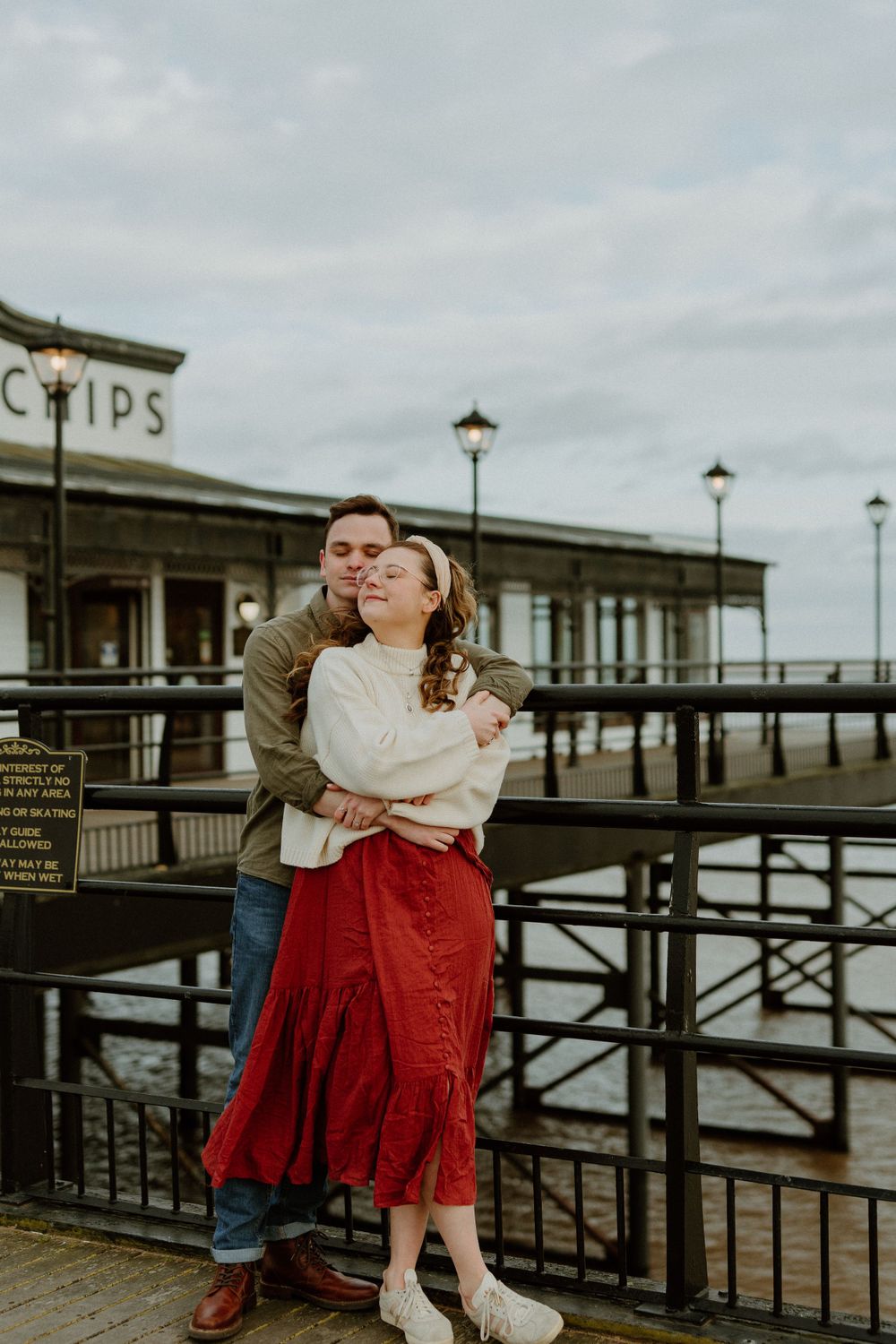 A couple embraces on a seaside pier with vintage lamp posts and railings on a cloudy day.