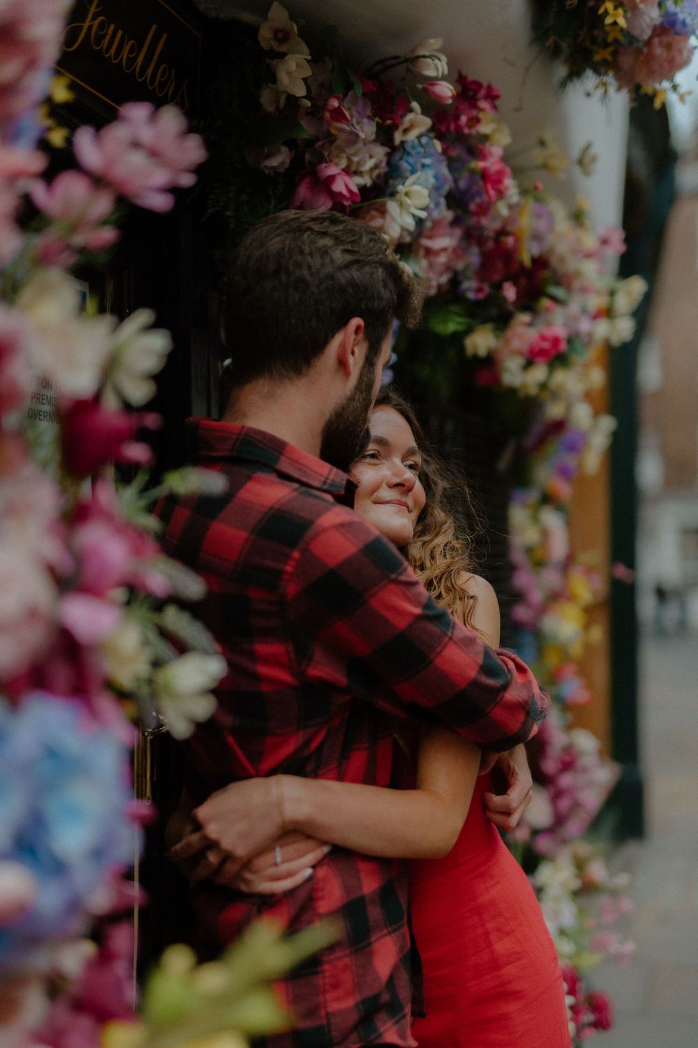 A couple embraces in front of a wall decorated with colorful pink and blue flowers.