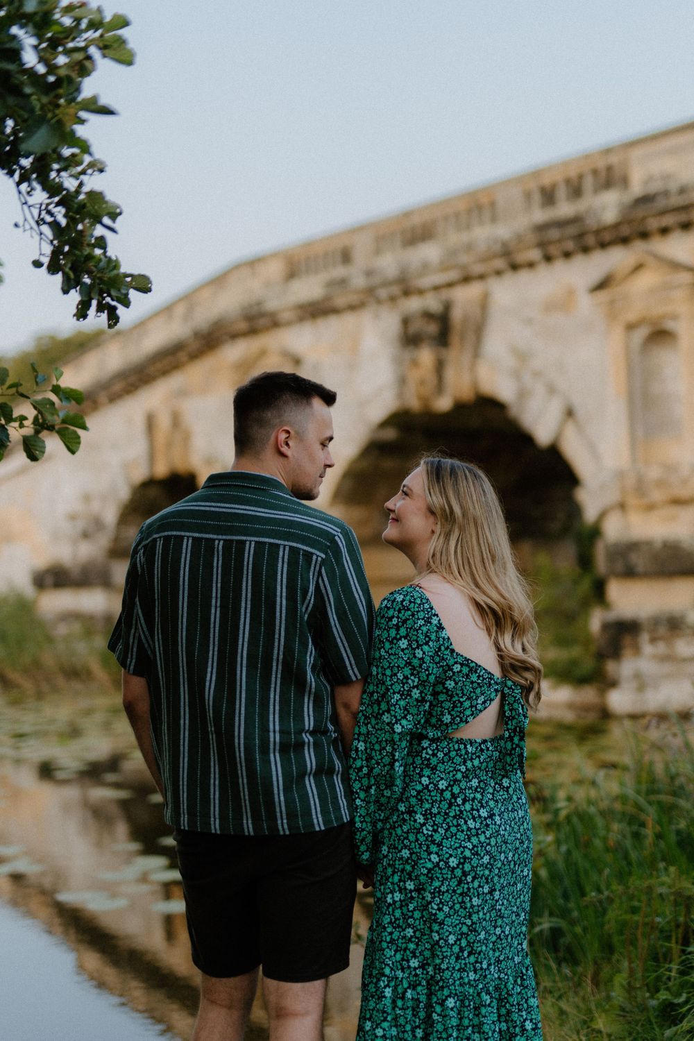 Couple in coordinated outfits standing by stone bridge and water reflection during golden hour photoshoot.