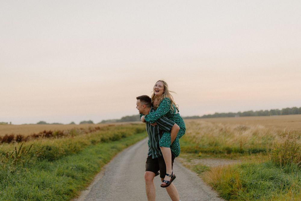 Two people share a playful piggyback ride along a country path at sunset surrounded by green fields.