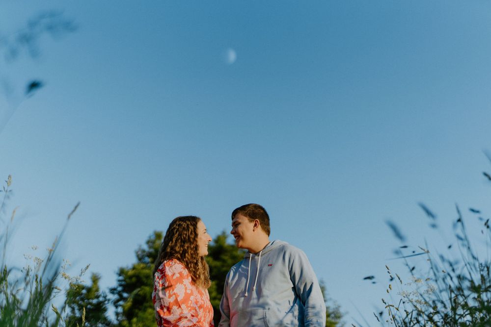 A couple stands close together against a blue sky with tall grass and trees in the background during golden hour.