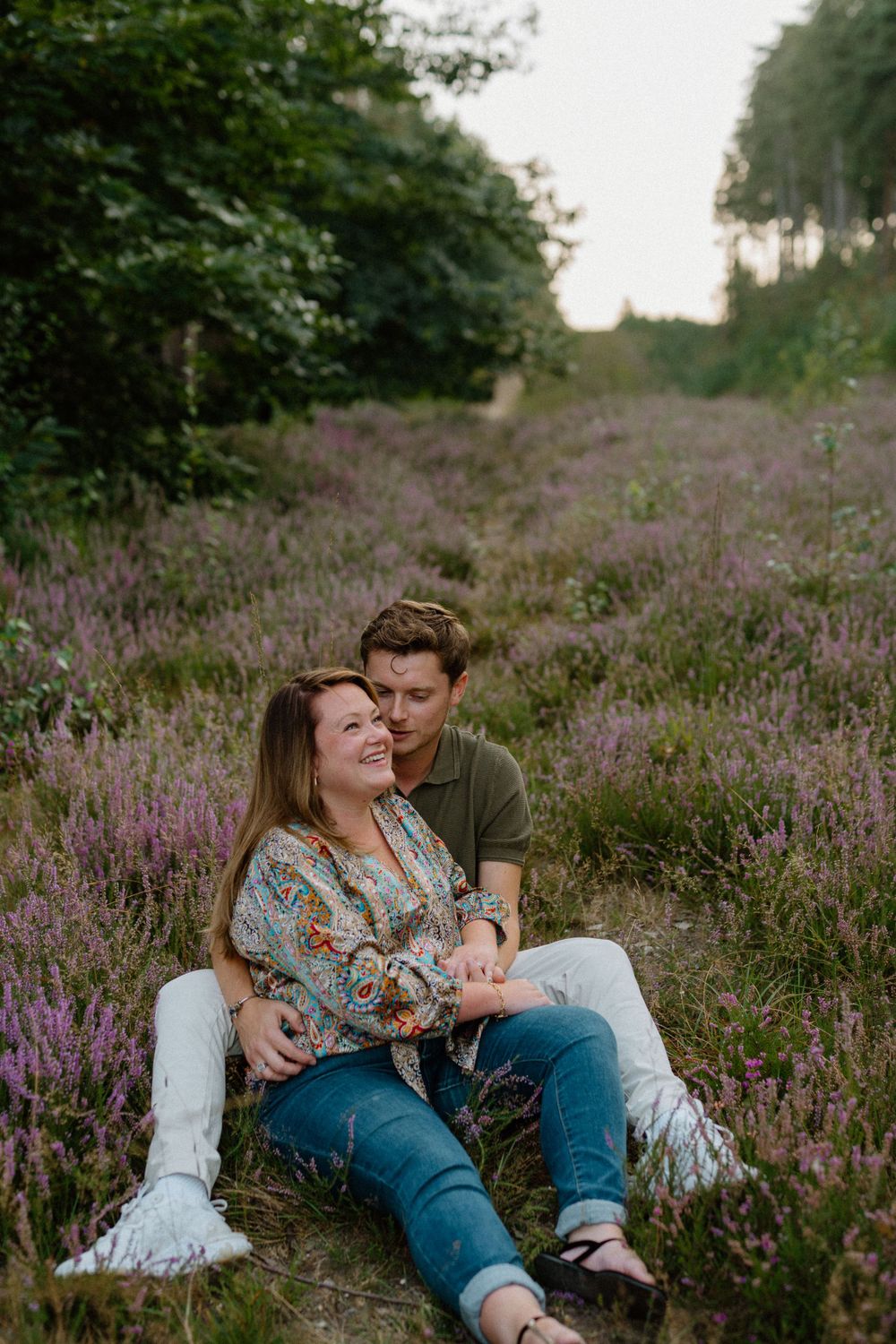 Couple sitting together in a blooming purple heather field at sunset surrounded by trees.