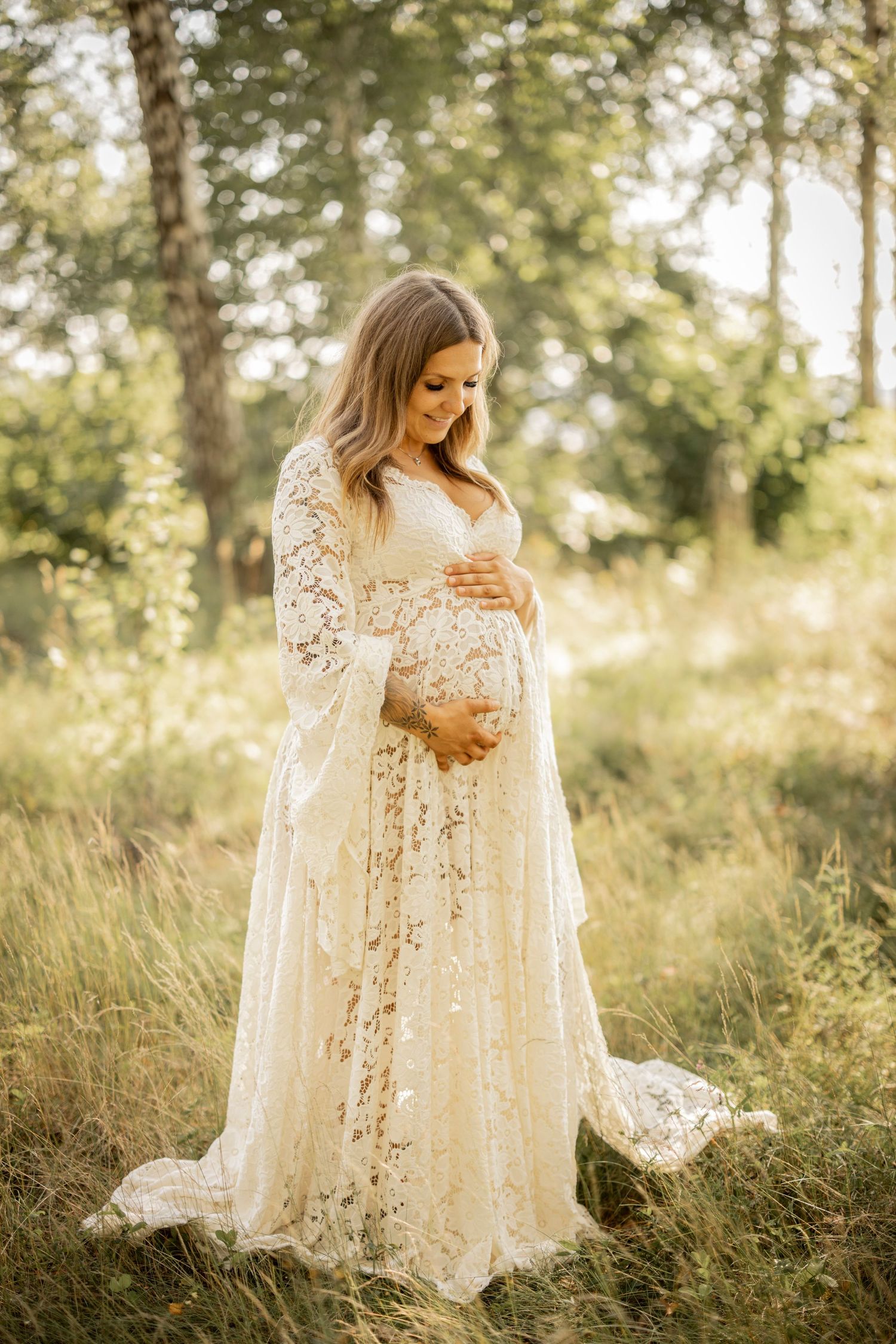 Expectant mother in flowing white lace maternity gown stands in sunlit meadow during golden hour photography session.