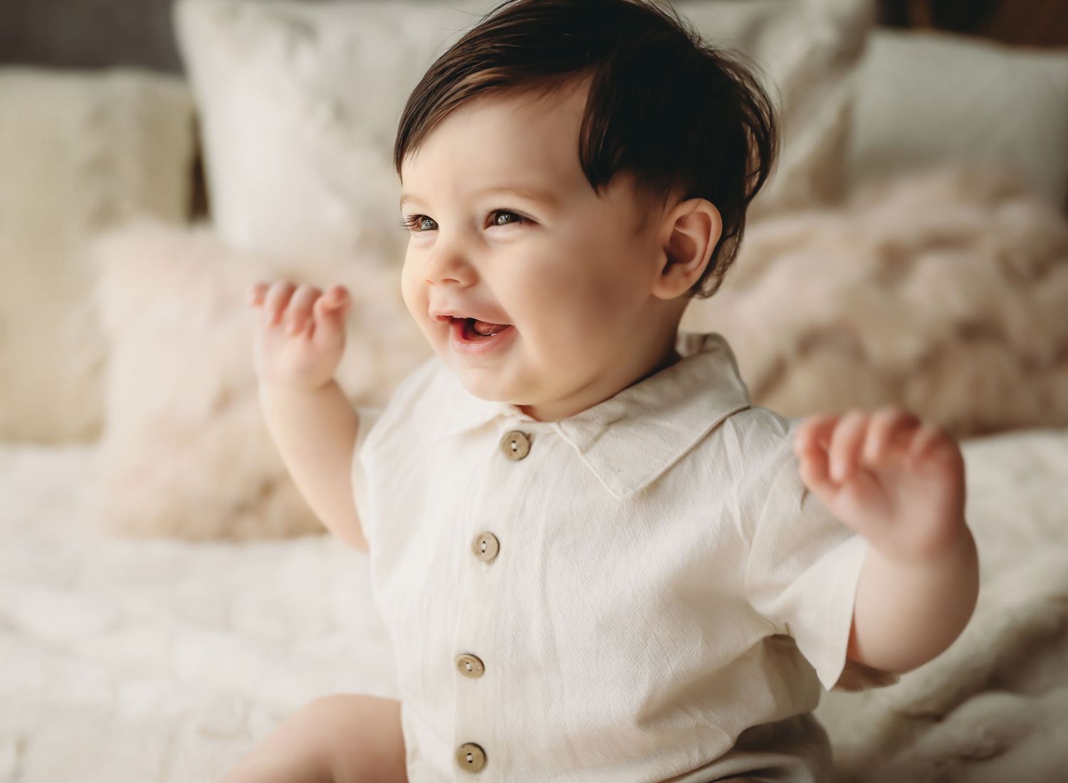 A baby in a blue dress sits on fluffy white fabric while smiling during a photo session.